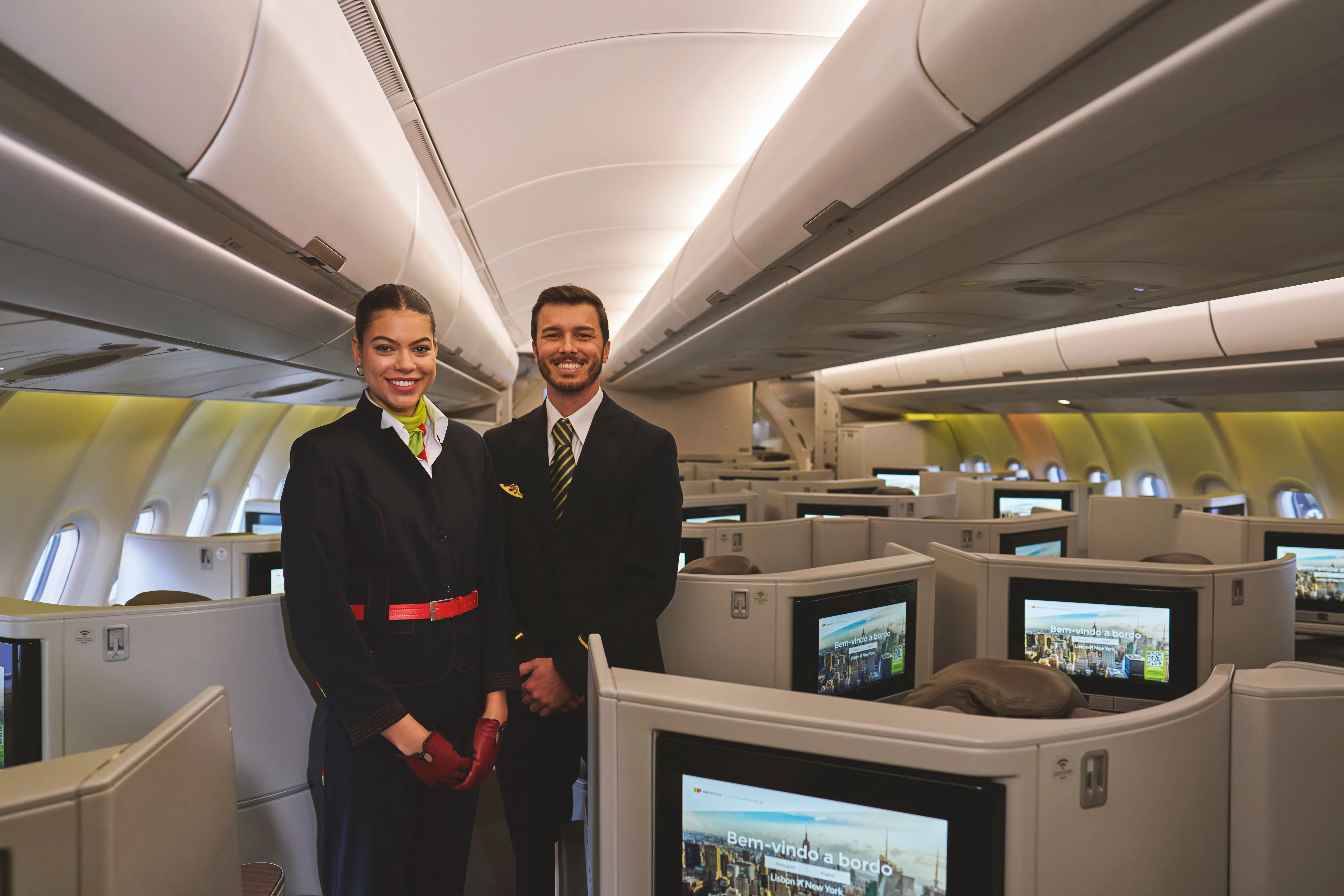 O interior de uma cabina de Classe Executiva de um avião da TAP. No centro da fotografia estão dois tripulantes de cabina, um homem e uma mulher, ambos vestidos com uniformes da TAP Air Portugal. São também visíveis vários assentos individuais com divisórias, cada um equipado com um ecrã de entretenimento.