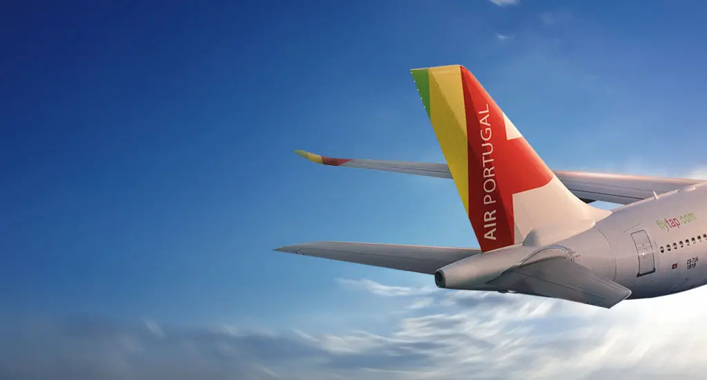 A parte traseira de um avião Airbus A330-900neo da TAP Air Portugal a voar acima das nuvens, com o céu azul como pano de fundo. O destaque vai para a cauda da aeronave, que exibe as cores verde, vermelho e amarelo, típicas da TAP, e a inscrição "AIR PORTUGAL" em letras brancas. Também é visível parte da fuselagem, onde se pode ler o endereço do website "flytap.com" em letras verdes e vermelhas.