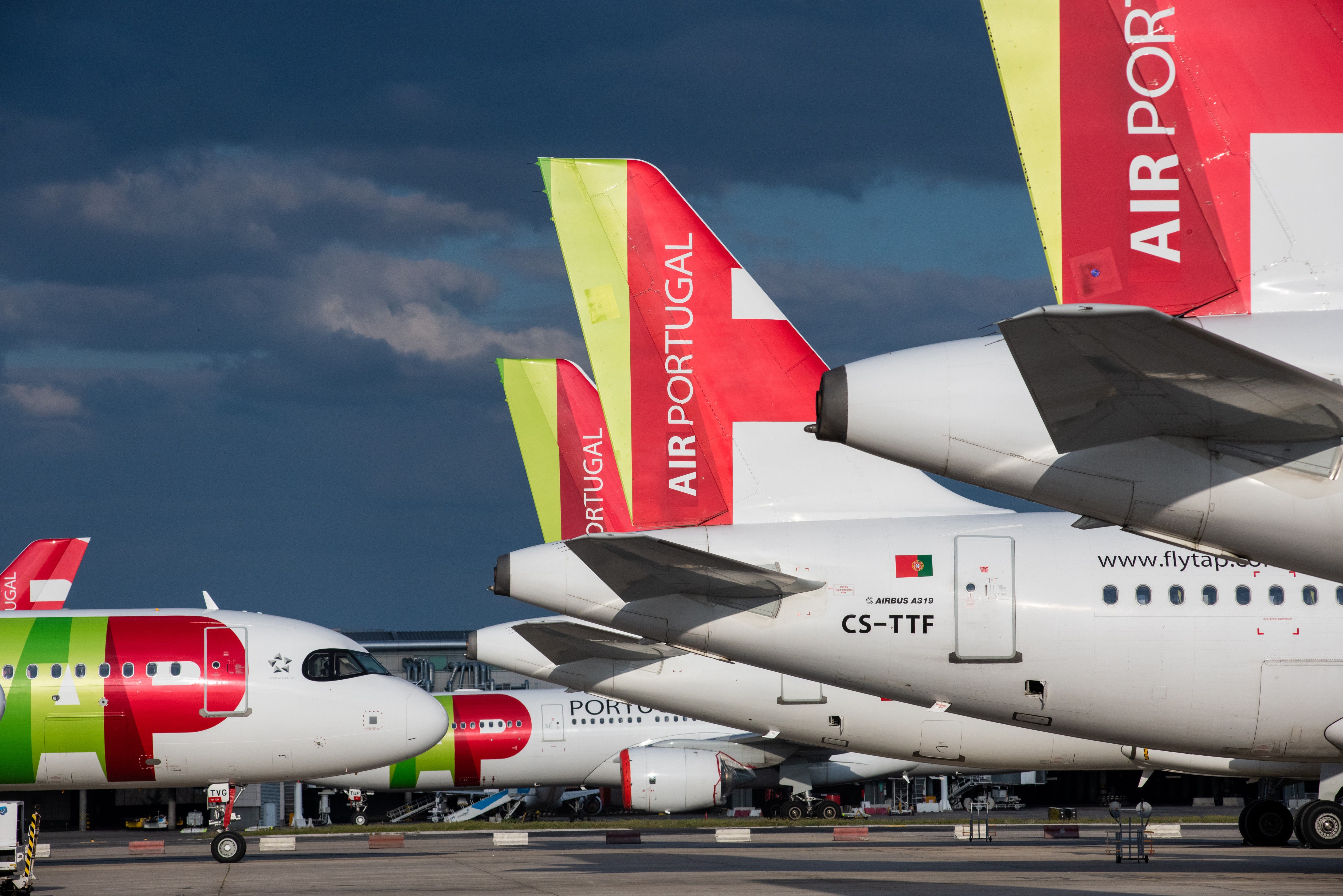 Vários aviões da TAP Air Portugal estacionados lado a lado num aeroporto. As caudas das aeronaves destacam-se com as cores verde, vermelho e branco, e a inscrição “AIR PORTUGAL”. Em primeiro plano, vê-se um Airbus A321, identificado pela matrícula CS-TTF e pelo nome do modelo junto à porta. O céu está parcialmente nublado.