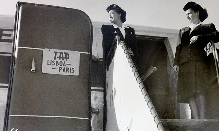 Imagem a preto e branco mostra duas assistentes de bordo da TAP no topo de uma escada de embarque, junto à porta de um avião. Na porta está afixada uma placa com a inscrição “TAP LISBOA - PARIS”, indicando o destino do voo. As assistentes envergam uniformes clássicos, compostos por casaco, saia e chapéu.